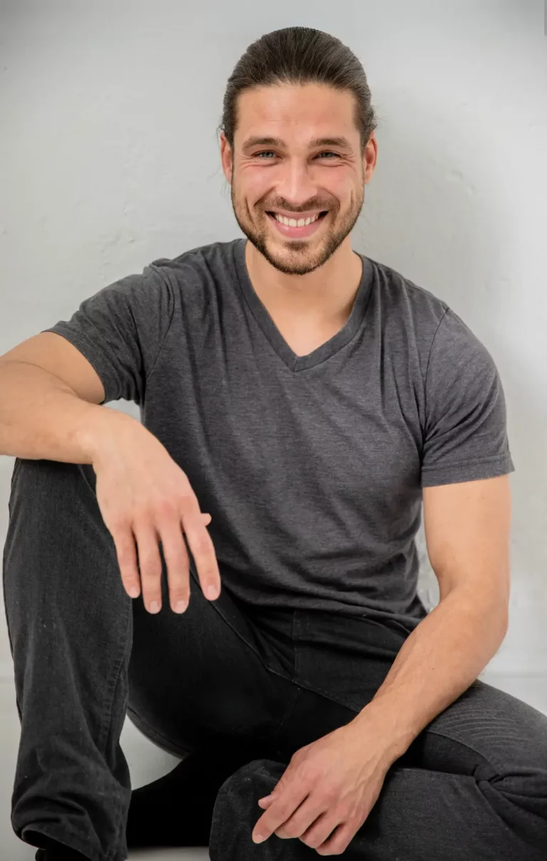 Portrait eines lächelnden Mannes in dunklem T-Shirt, der entspannt vor einer hellen Wand sitzt – freundliche, authentische Ausstrahlung.